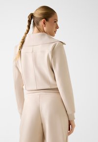 Veste de sport beige avec un large col, des poignets et un ourlet côtelés. Texture douce avec des détails de coutures visibles. Cheveux coiffés en tresse.