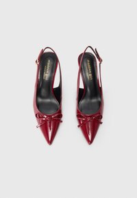 Punaiset lakatut slingback-korkokengät, joissa on teräväkärkinen malli, etuosassa on rusettiyksityiskohta ja kultainen solki nilkkahihnassa.