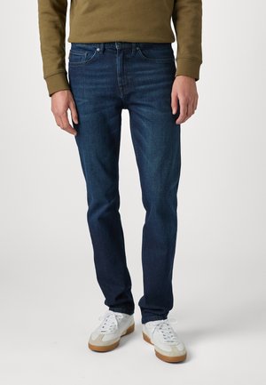 Sötétkék, slim fit farmernadrág, finom kopásokkal, öt zsebes kialakítással, fehér tornacipőkkel párosítva.