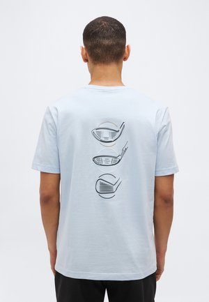 Muž v světle modrém tričku s třemi vertikálně vyobrazenými obrysy hlav golfových holí na zádech, stojící na jednobarevném bílém pozadí.