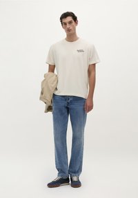 Beige bomulds t-shirt med et lille sort logo, parret med lyseblå lige ben jeans og mørkeblå sneakers. Modellen holder en lys jakke.
