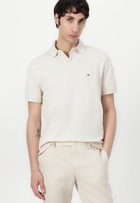 Beige poloshirt met een klassieke kraag, korte mouwen en een klein geborduurd logo op de borst, gecombineerd met lichtgekleurde broeken.