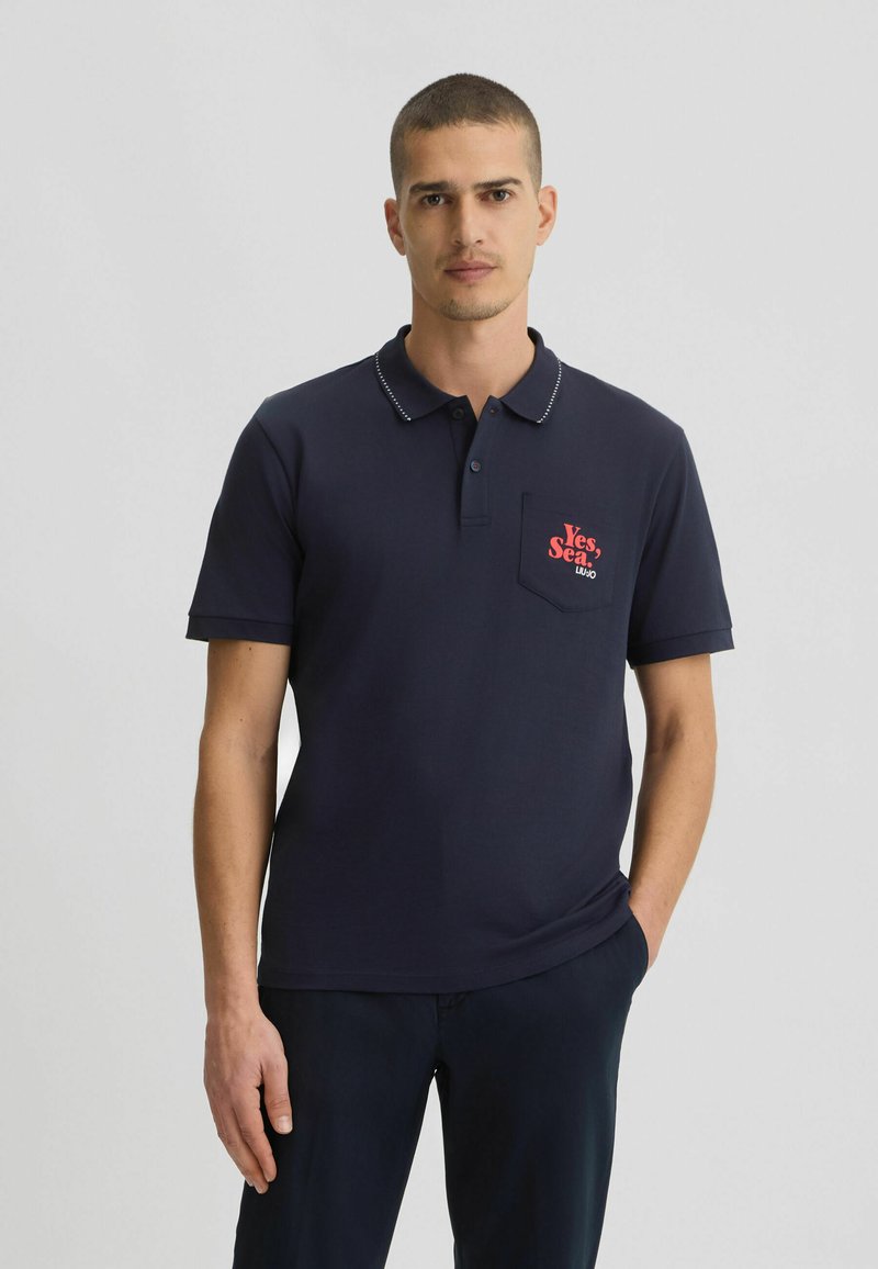 Camiseta tipo polo navy con cuello, que presenta un logotipo rojo en el bolsillo. Fabricada en una tela suave, de mangas cortas y con un sutil ribete estampado.