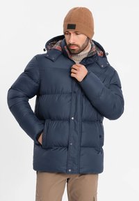 Giubbotto imbottito blu navy con cappuccio e tasche frontali, con fodera a quadri e bottoni a pressione. Cuffia lavorata in lana di colore marrone.