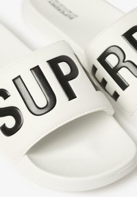 Hvide skridsikre sandaler med en tekstureret sål og en bred rem med fede sorte bogstaver, der staver "SUPER." Glat syntetisk materiale.