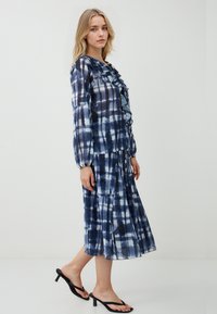 Donna che indossa una blusa a maniche lunghe blu a fantasia con volant e una gonna midi plissettata abbinata, abbinata a sandali con tacco neri.