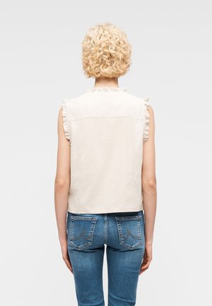 Personne blonde aux cheveux bouclés portant un gilet beige sans manches avec des bords à volants et un jean bleu en denim, debout dos à la caméra sur un fond uni.
