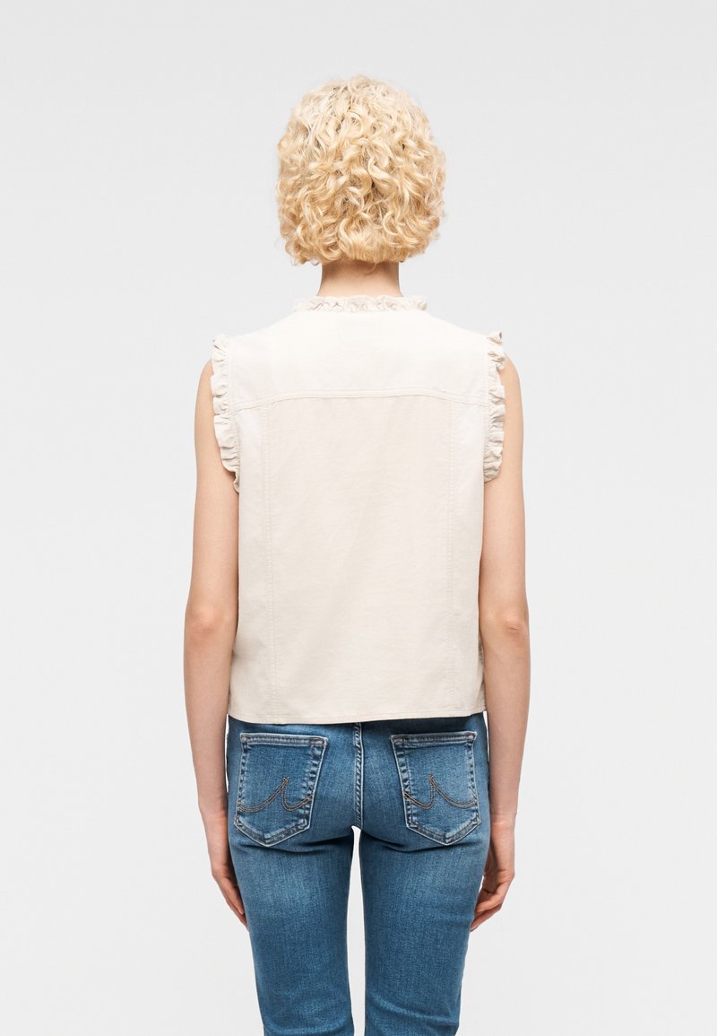 Personne blonde aux cheveux bouclés portant un gilet beige sans manches avec des bords à volants et un jean bleu en denim, debout dos à la caméra sur un fond uni.