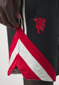 Los pantalones cortos deportivos negros presentan acentos diagonales rojos y blancos, paneles laterales a rayas y un logo bordado en rojo de una figura sosteniendo un tridente.