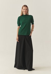 Zielony top z krótkim rękawem i wysokim dekoltem, zestawiony z czarnymi szerokimi spodniami. Gładka tekstura z subtelnym połyskiem. Złota bransoleta na mankiecie jako akcent.