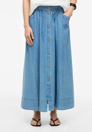 Femme portant une longue jupe en denim bleu avec des boutons à l’avant, des poches latérales, un haut blanc, un bracelet noir et des sandales noires.