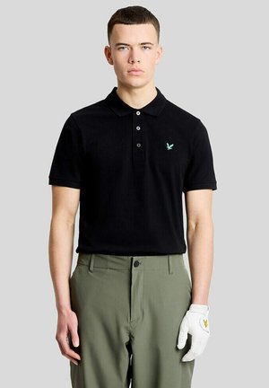 Ung mand iført en sort poloshirt, grønne bukser og en hvid golfhandske på venstre hånd mod en ensfarvet baggrund.
