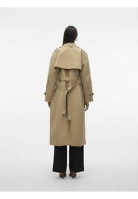 Beige trenchcoat med belted midje, stor krage og skulderepauleter. Har en bakflap og en avslappet passform. Kombinert med svarte bukser.