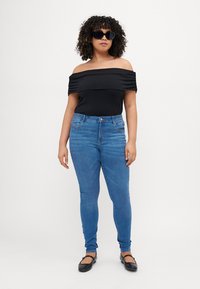 Black off-shoulder top with gathered detail, paired with blue skinny jeans. Model wears black sunglasses and flat black shoes.