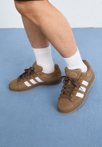 Brown suede sneakers feature three white stripes, brown laces, and a rubber sole. Worn with white socks, set against a blue background.