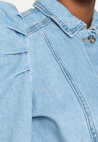 Veste en denim bleu clair avec détails aux épaules bouffantes, col classique et fermeture à boutons argentés avec surpiqûres contrastées.