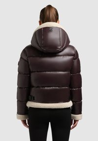 Giacca piumino marrone scuro con un ampio cappuccio, foderata in shearling crema al colletto e ai polsini, con sezioni trapuntate orizzontali e vita regolabile.