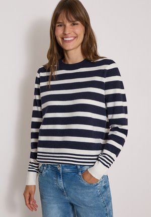 Femme souriante portant un pull à rayures horizontales bleu marine et blanc et un jean bleu, une main dans la poche, sur fond uni.