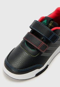 Chaussures synthétiques noires avec deux sangles ajustables en Velcro, surface texturée, doublure intérieure rouge et détails de couture colorés.