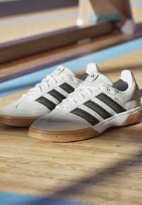 Weiße und schwarze Adidas Spezialist Sneaker mit Gummisohlen, auf poliertem Holzboden nahe einem blau gestreiften Sportplatz platziert.