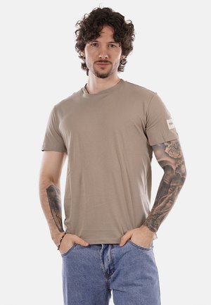 Uomo con capelli ricci, t-shirt beige, jeans blu e tatuaggi dettagliati sulla manica, in piedi con le mani nelle tasche su sfondo bianco.