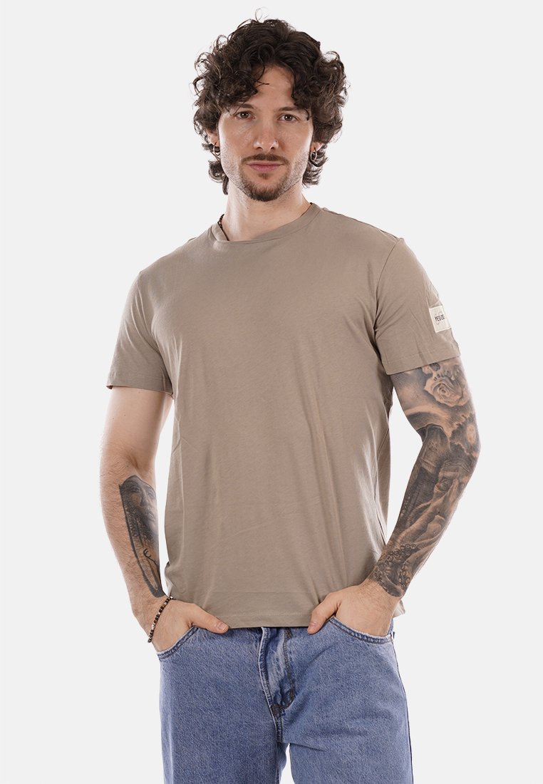 Homme aux cheveux bouclés, portant un t-shirt beige, un jean bleu et des tatouages détaillés sur les manches, debout les mains dans les poches devant un fond blanc.