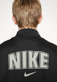 Giacca da atleta nera con colletto alto, caratterizzata da grande scritta bianca "NIKE" e piccolo logo swoosh sul retro. Tessuto dalla texture liscia.