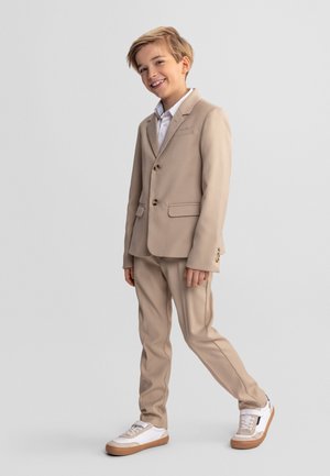 Uśmiechnięty chłopiec stoi, ubrany w beżowy garnitur z białą koszulą i białymi trampkami, pozując na jednolitym jasnoszarym tle.