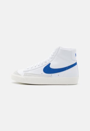 Biały, wysokogatunkowy sneaker z niebieskim logo Nike swosh i białymi sznurówkami, widziany od wewnętrznej strony na jednolitym tle.