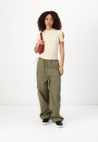Beige kortermet topp med skulderutskjæringer, kombinert med olivengrønne cargo-bukser og svarte sneakers. Modellen holder en rød crossover-bag.