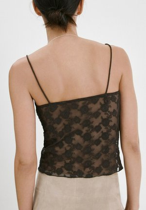 Žena s tamnom kosom u niskoj punđi, odjevena u crnu cvjetnu čipkastu majicu bez rukava s tankim naramenicama i bež donji dio, prikazana s leđa.