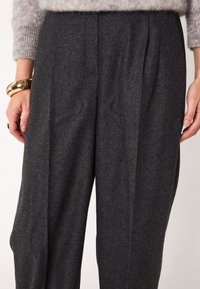 Pantaloni larghi grigio scuro realizzati in un tessuto morbido e testurizzato, con pieghe frontali e una vestibilità comoda, indossati con un bracciale dorato.