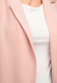 Lys pink blazer med en glat tekstur og notch-lapel, båret over en hvid T-shirt med rund hals. Minimalistisk design og blødt stof.