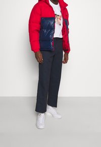 Röd och marinblå vadderad jacka med huva, vit t-shirt med tryck, mörkblå byxor och vita sneakers. Slät textur och färgblockdesign.