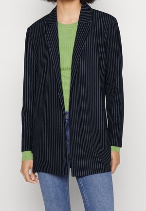 Person wearing a black pinstripe blazer over a green ribbed sweater and blue jeans, shown from neck to mid-thigh.