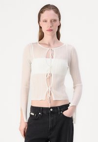 Hvit ribbestrikket genser med lange ermer, rund hals og frontknyting. Bært over svarte høyhalsede jeans med åpninger på ermene.