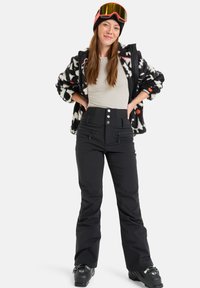 Zwarte waterdichte skibroeken met knoopsluiting, gecombineerd met een gestreept zwart-wit fleecjacket. Model draagt skibril en een beanie.