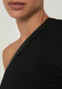 Detailný záber na osobu v čiernom topu na jednom ramene so zdobeným čiernym perlovým lemom pozdĺž výstrihu.