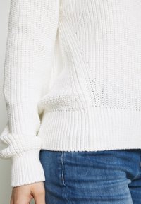 Persona con un suéter de punto grueso blanco con puños y dobladillo acanalados, combinado con jeans de mezclilla azul, mano relajada al costado.