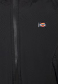 Black jacket with a smooth texture, featuring a central zipper and a small Dickies logo patch on the upper left side. No visible patterns.
