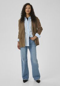 Brun kordfløjl blazer over en lyseblå skjorte med knapper, parret med lys blå flared jeans og sorte spidse sko, mod en ensfarvet baggrund.
