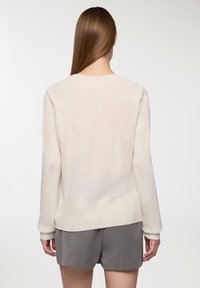 Kremowy sweter robiony na drutach z ribbowym wykończeniem u dołu i na mankietach, z luźnym krojem i teksturowanym wzorem, noszony na szaro-białe szorty.