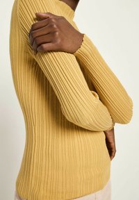 Eine dunkelhäutige Person trägt einen gerippten, senfgelben Langarmpullover, wobei eine Hand auf dem gegenüberliegenden Oberarm ruht.