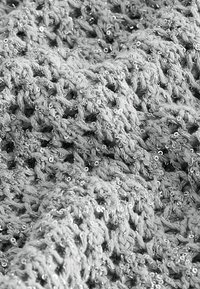 Zbliżenie na grubą dzianinę, wplataną w małe błyszczące cekiny, podkreślającą teksturowane pętelki włókna i odblaskowe detale.