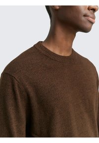 Brauner Strickpullover mit Rundhalsausschnitt und strukturiertem Stoff, der ein dezentes Muster hervorhebt. Nahaufnahme des Kragens und der Schulter.