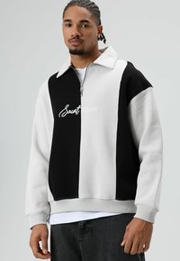 Zwart-witte sweater met ritssluiting en kraag, met "Saintessence" geborduurd aan de voorkant en geribbelde manchetten.