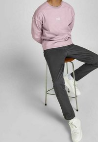 Ljusrosa sweatshirt med rundad hals och vit logotyp, tillsammans med mörkgrå slim-fit-byxor och vita sneakers, sittande på en metallpall.