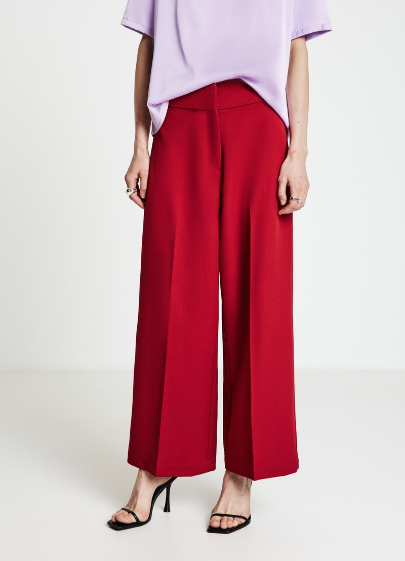 Pantalon rouge à jambes larges en tissu lisse, doté d'une taille haute et d'une silhouette structurée. Associé à un haut violet clair et des talons noirs.