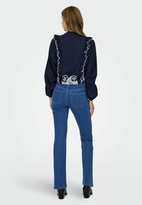 Blusa azul marino con mangas con volantes y detalle de encaje en el dobladillo, combinada con vaqueros ajustados azules y zapatos de tacón negros.