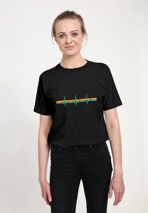 Kvinde iført en sort t-shirt med en grøn hjerterytmeline over en vandret regnbuestribe på brystet, stående mod en hvid baggrund.